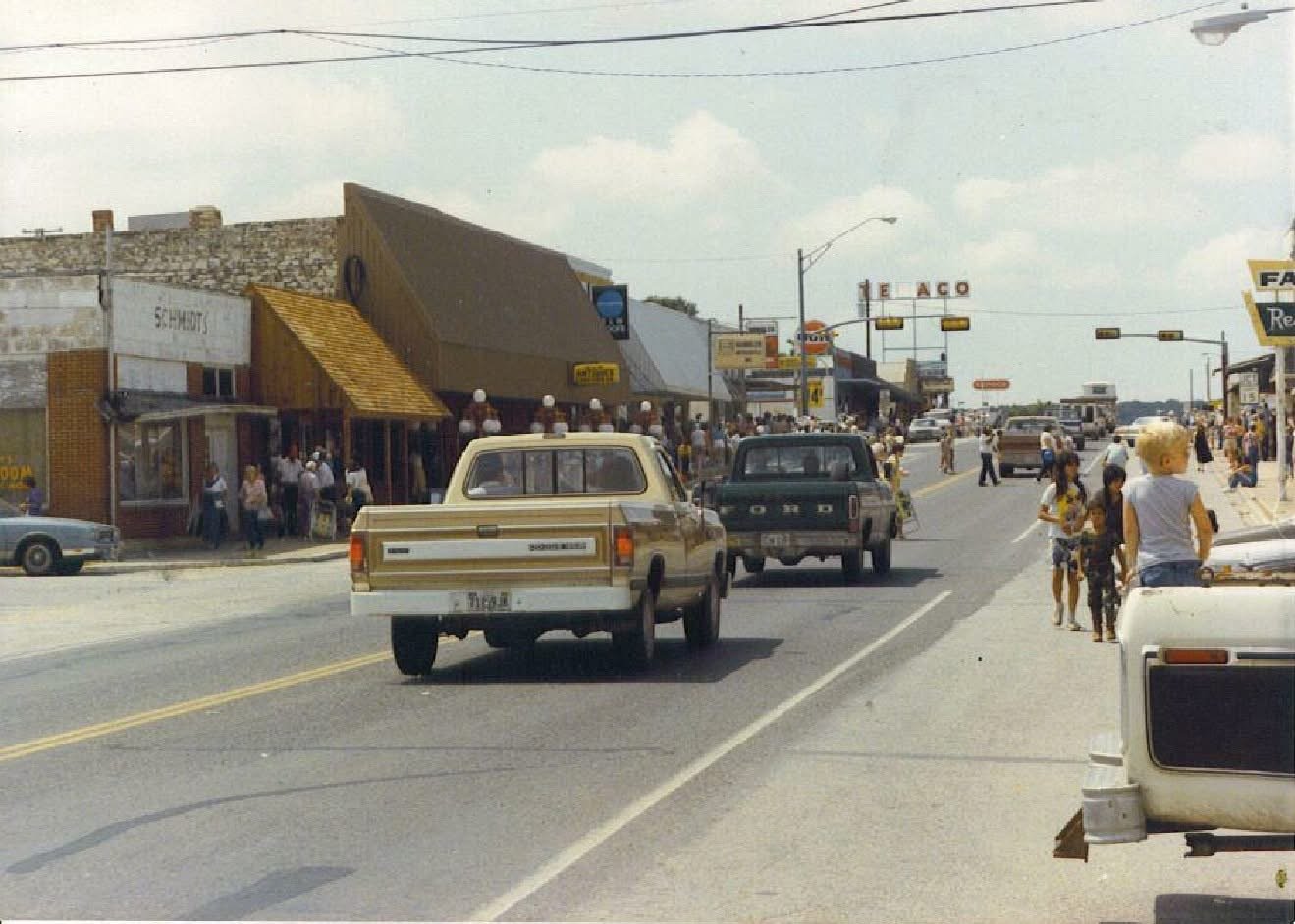 Vintage photograph of pickup trucks driving down a small-town main street with storefronts and pedestrians.