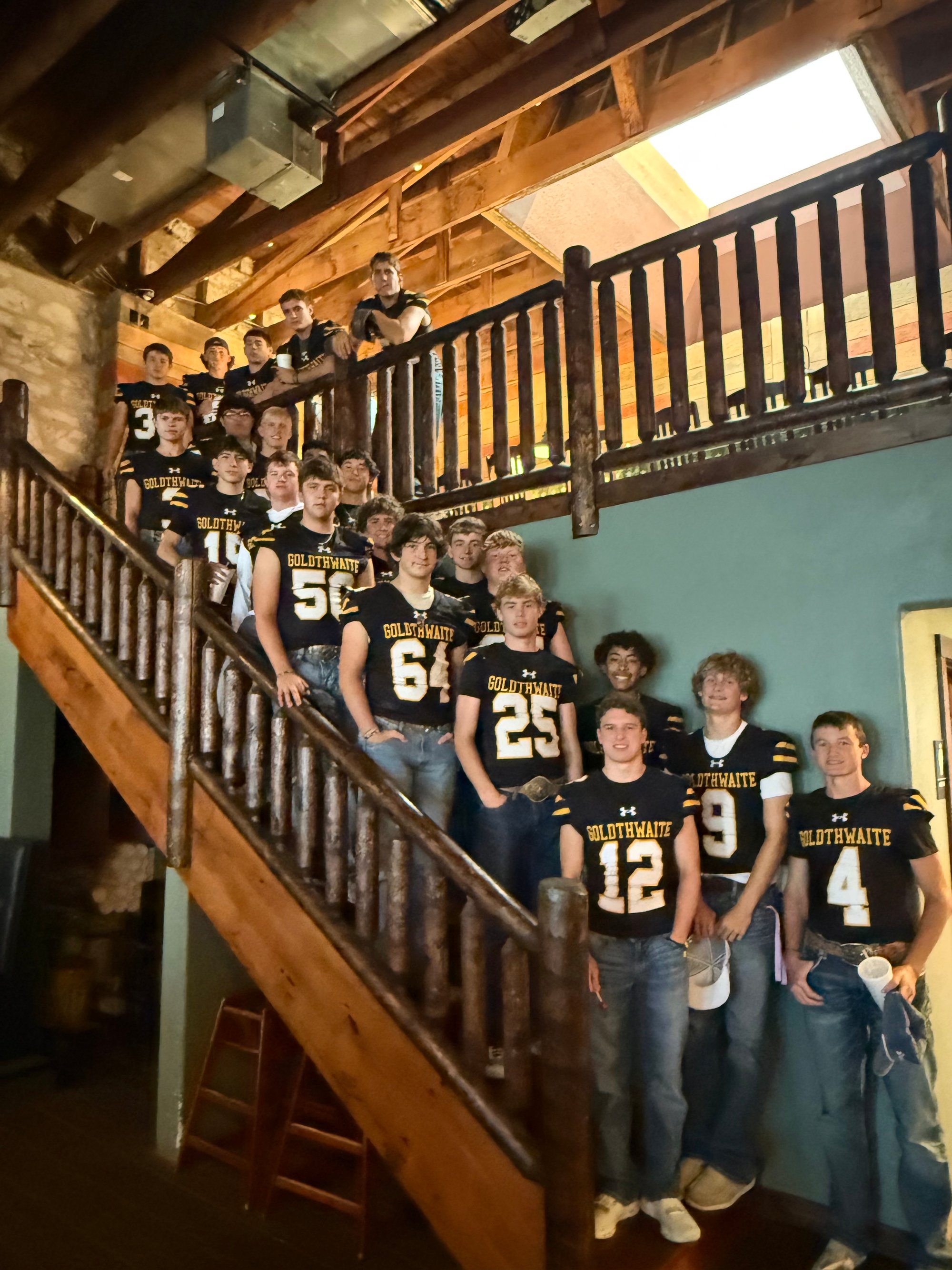 High school football players wearing black and yellow jerseys pose on a wooden staircase inside a restaurant.